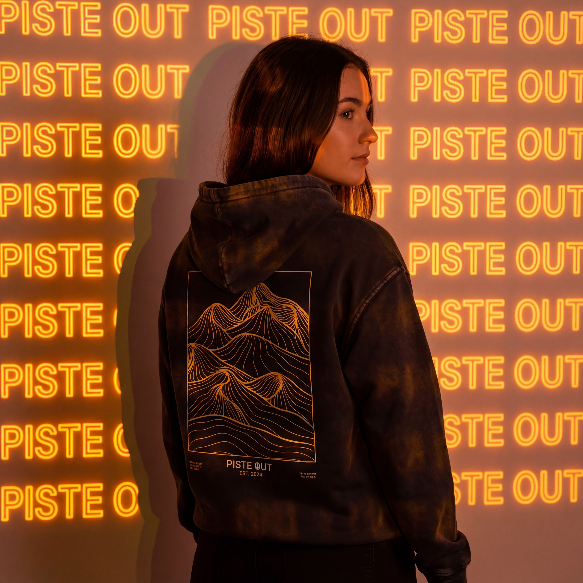 Model shot of the Black Diamond backcountry veins hoodie showing the back gold line work graphic. Piste Out is projected in orange neon on the wall.