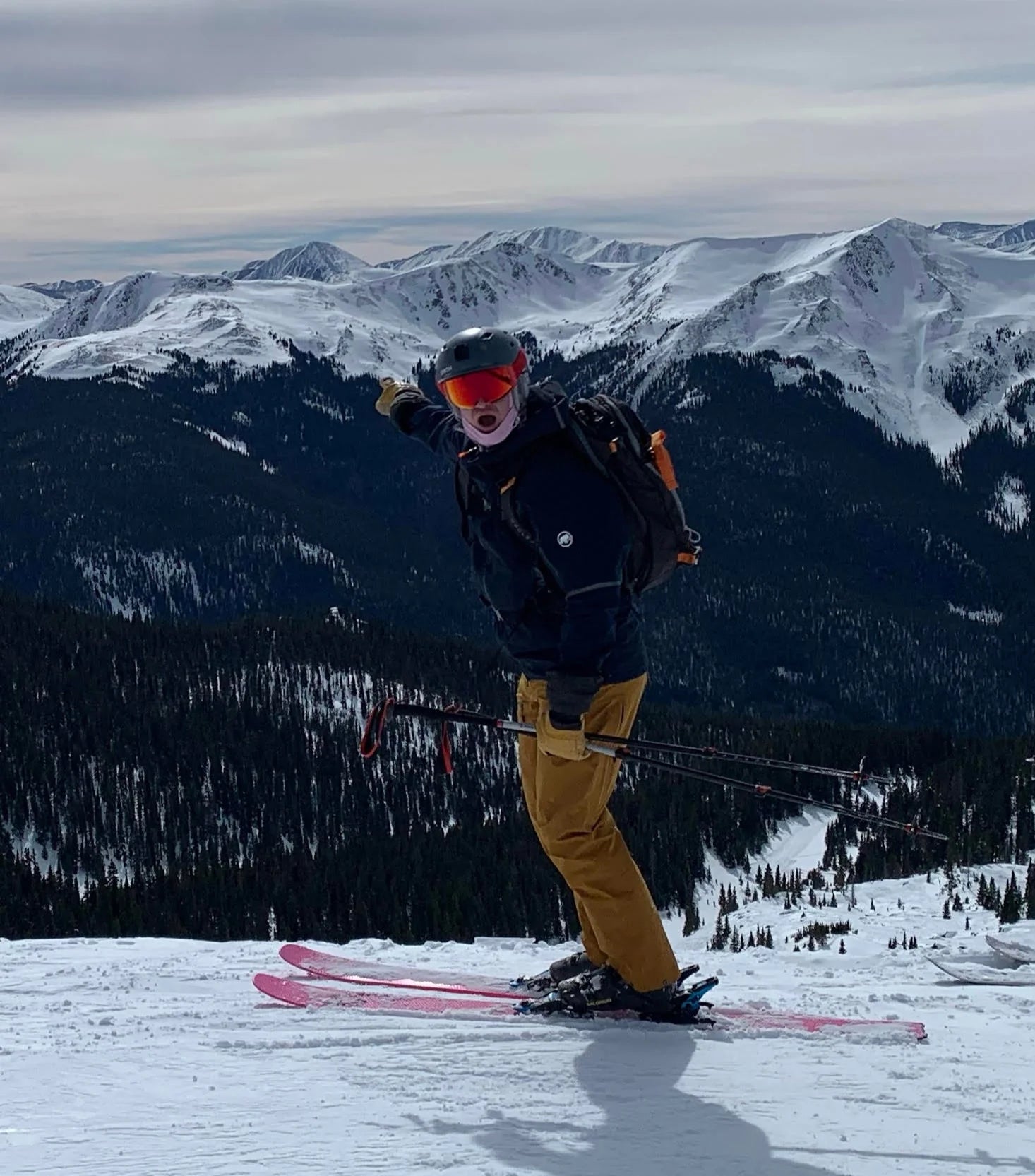 Piste Out founder standing on a snowy ridgeline pointing toward a snowcapped mountain range while backcountry skiing