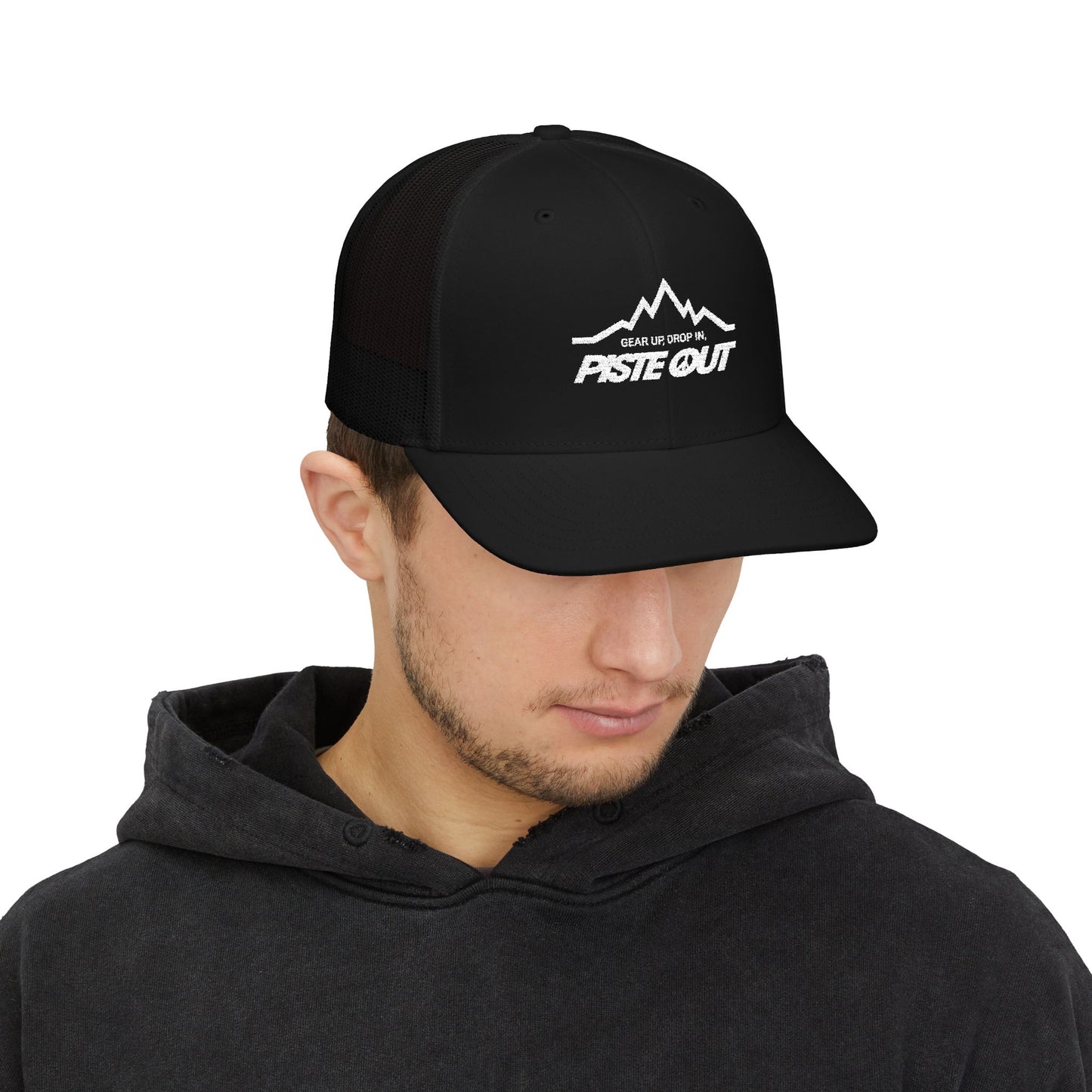 Black trucker hat worn by model, with white Piste Out mountain logo and slogan “Gear Up. Drop In.” embroidered on front