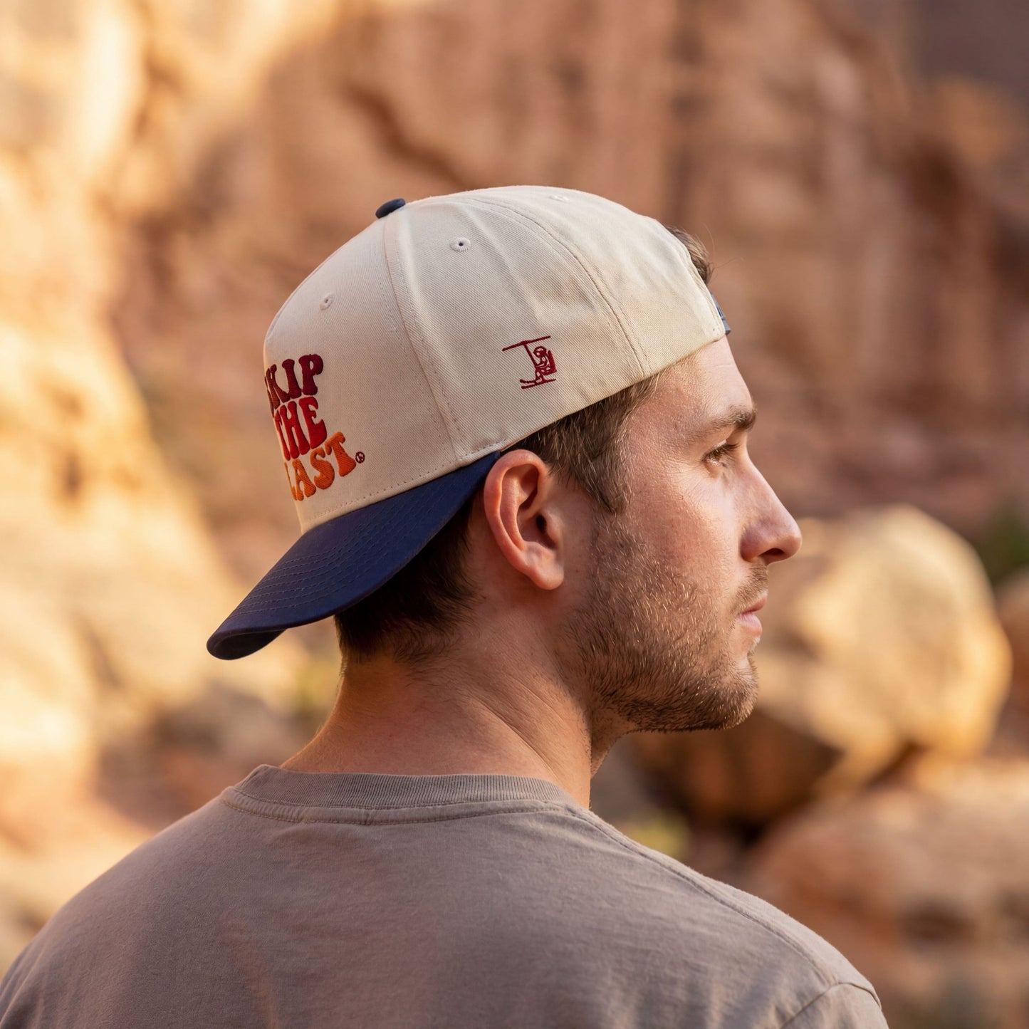 Angled view of the Skip the Last hat in Blue Square, with cream crown, blue brim, and side skier lift embroidery, worn on a male model.