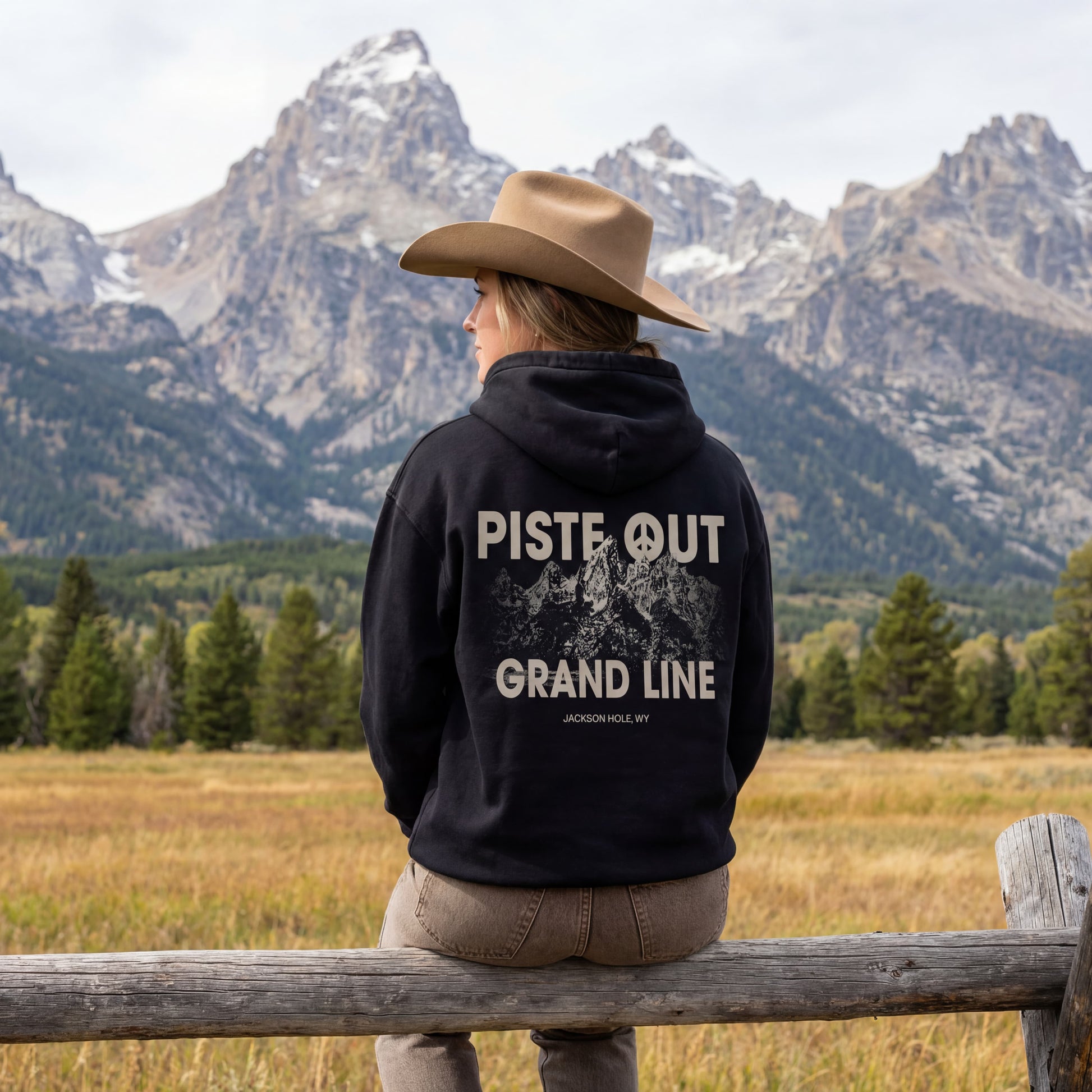 Model shot of female model wearing the grandlines hoodie and cowboy hat, sitting on wooden fence in front of the Tetons. 