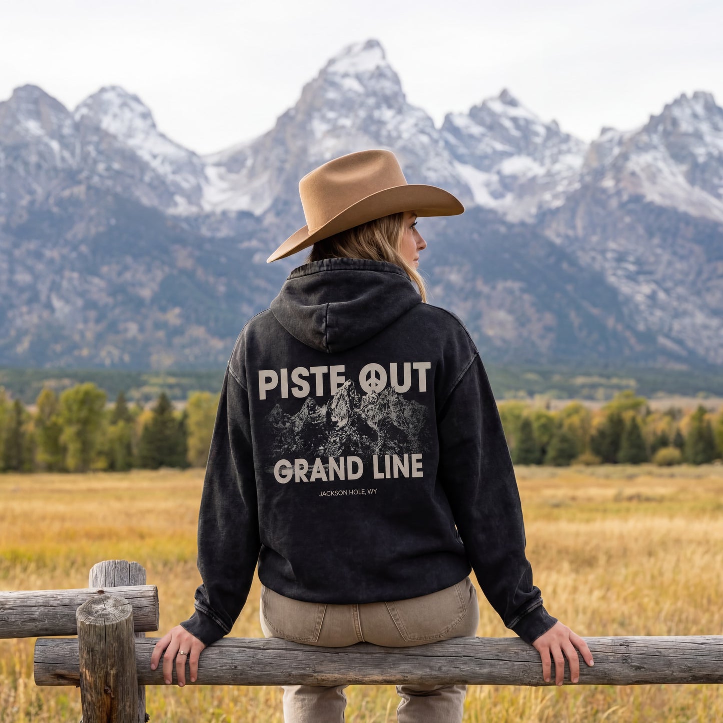 Model shot of female model wearing the grandlines hoodie and cowboy hat, sitting on wooden fence in front of the Tetons. 