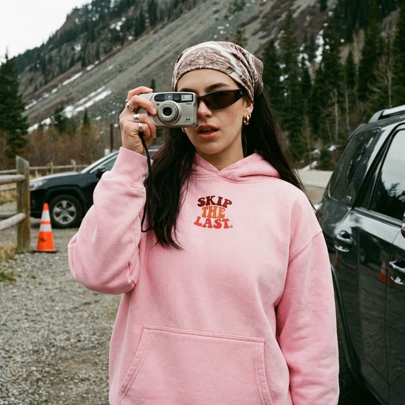 Women wearing the Hoodie in Pink, front view with chest logo with a point and shoot camera in from of mountains. 