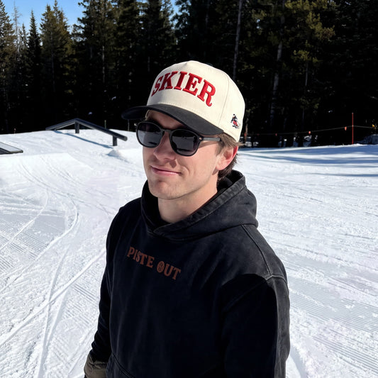 A man standing on a snowy mountain slope wearing a beige and black trucker hat with 'SKIER' in red, a black hoodie with 'PISTE OUT' in black, sunglasses, and gloves. Trees and ski slopes are visible in the background.