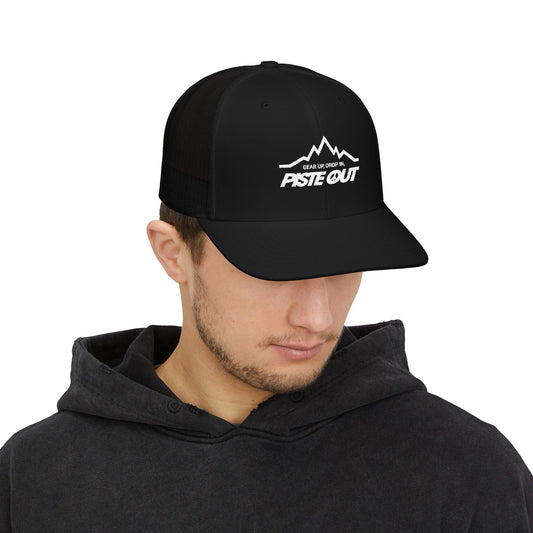 Black trucker hat worn by model, with white Piste Out mountain logo and slogan “Gear Up. Drop In.” embroidered on front
