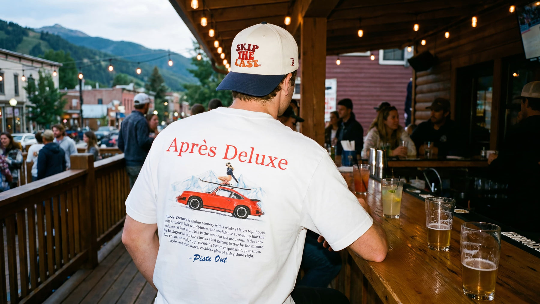 Man wearing Skip the Last hat and Après Deluxe tee at a mountain deck bar with Colorado mountain views